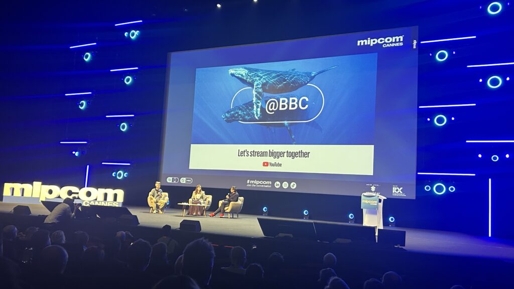 Panel discussion on stage at MIPCOM Cannes featuring BBC Studios during the YouTube keynote, with a large screen displaying BBC branding and a message about streaming, highlighting the company’s shift to social-first sales teams.