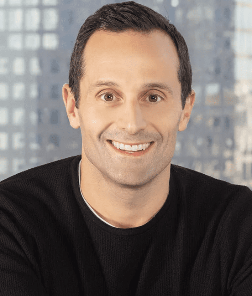 Portrait of a smiling man with short dark hair, wearing a black sweater, against a blurred urban background.