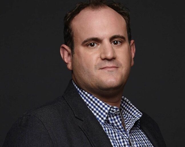 Professional headshot of a media and entertainment executive wearing a dark blazer and checkered shirt against a dark background.