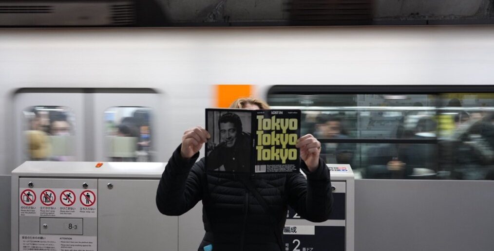 Person holding a copy of Lost in Tokyo on a Tokyo metro platform with a train passing in the background.